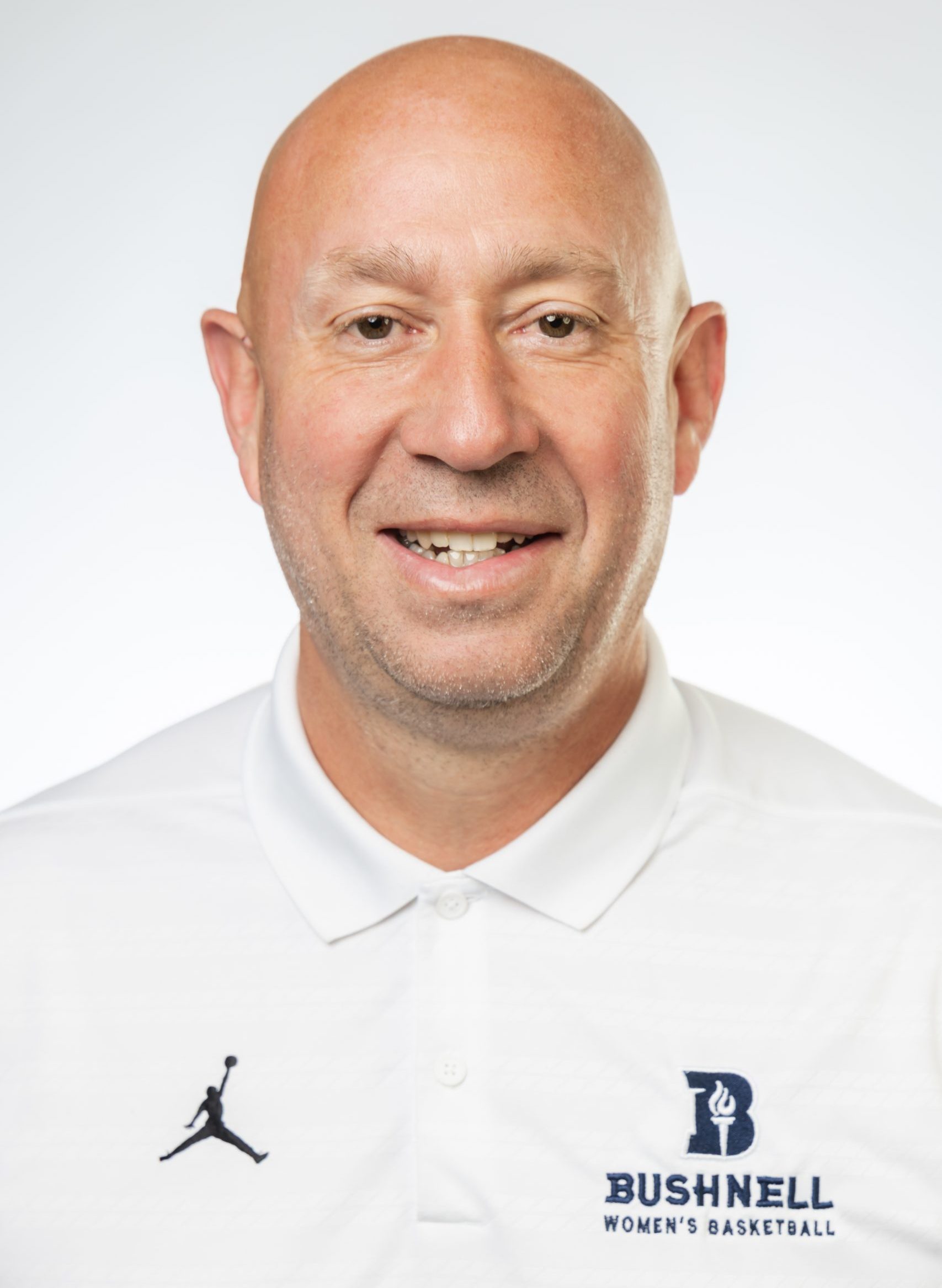 Headshot of a smiling bald man with light stubble, wearing a white Bushnell Women’s Basketball polo against a white background