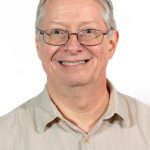 Headshot of a smiling older man with short gray hair and glasses, wearing a beige button-down shirt against a white background