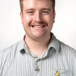 Headshot of a smiling man with short light brown hair and a mustache, wearing a light plaid button-down shirt and cross necklace against a white background