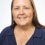 Headshot of a smiling woman with long straight brown hair, wearing a navy blouse and silver necklace against a white background