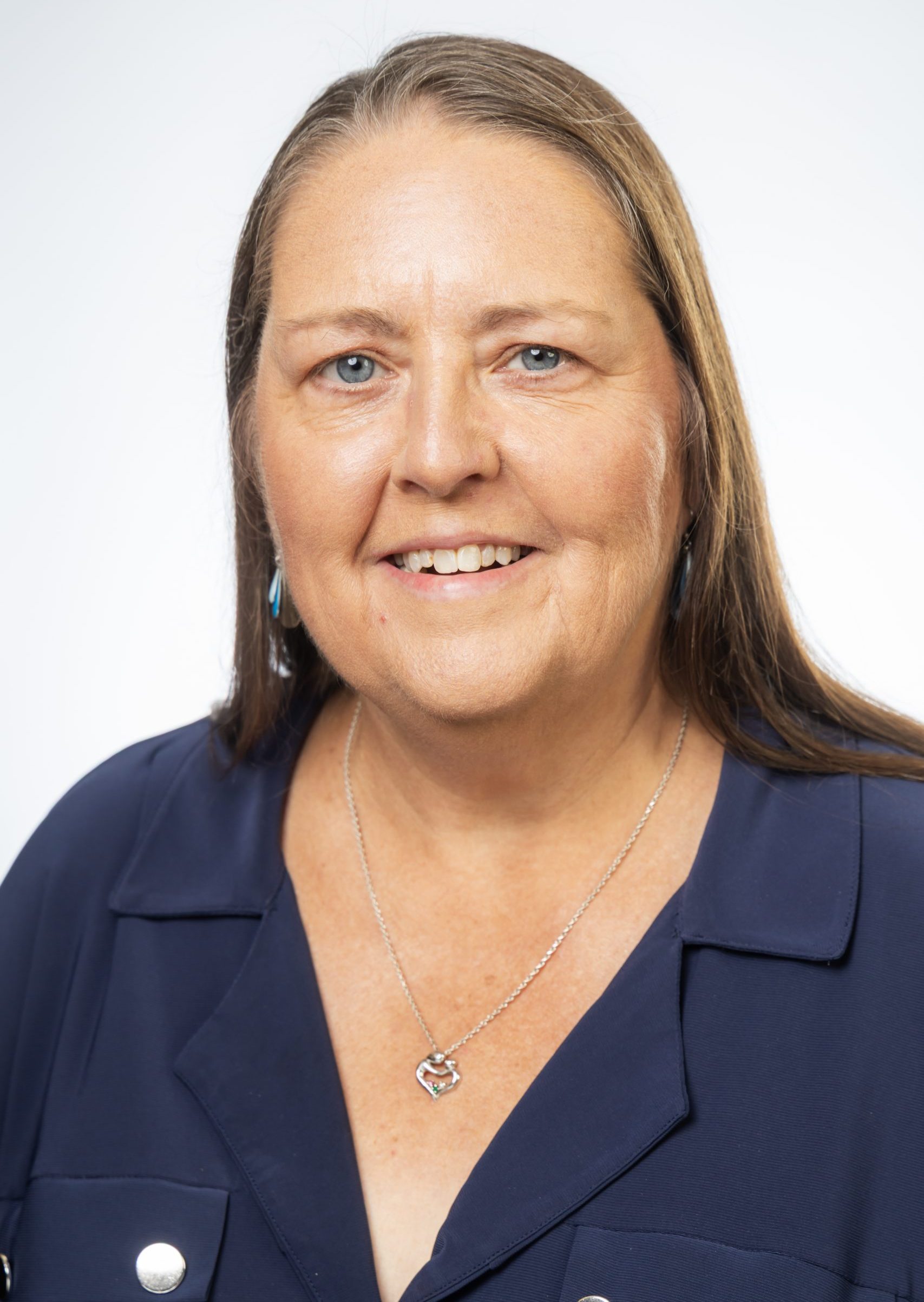 Headshot of a smiling woman with long straight brown hair, wearing a navy blouse and silver necklace against a white background