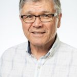 Headshot of a smiling older man with gray hair and glasses, wearing a light checkered button-down shirt against a white background
