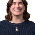 Headshot of a smiling woman with short wavy brown hair, wearing a navy sweater and pendant necklace against a white background