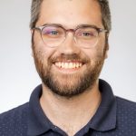 Headshot of a smiling man with short hair, glasses, and a beard, wearing a navy polo shirt against a white background