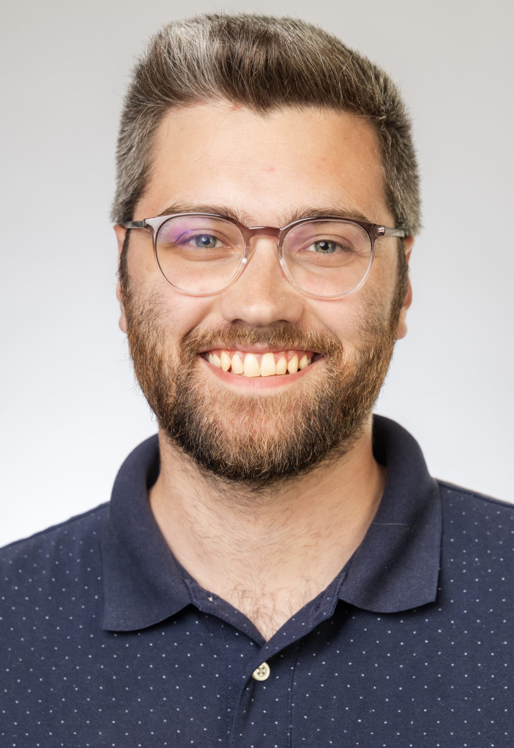 Headshot of a smiling man with short hair, glasses, and a beard, wearing a navy polo shirt against a white background