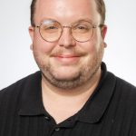 Headshot of a smiling man with short hair, glasses, and a beard, wearing a black polo shirt against a white background