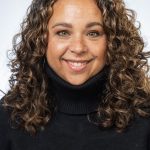 Headshot of a smiling woman with shoulder-length curly hair, wearing a black turtleneck sweater against a white background