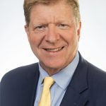 Headshot of a smiling man with light brown hair wearing a navy suit and yellow tie against a white background