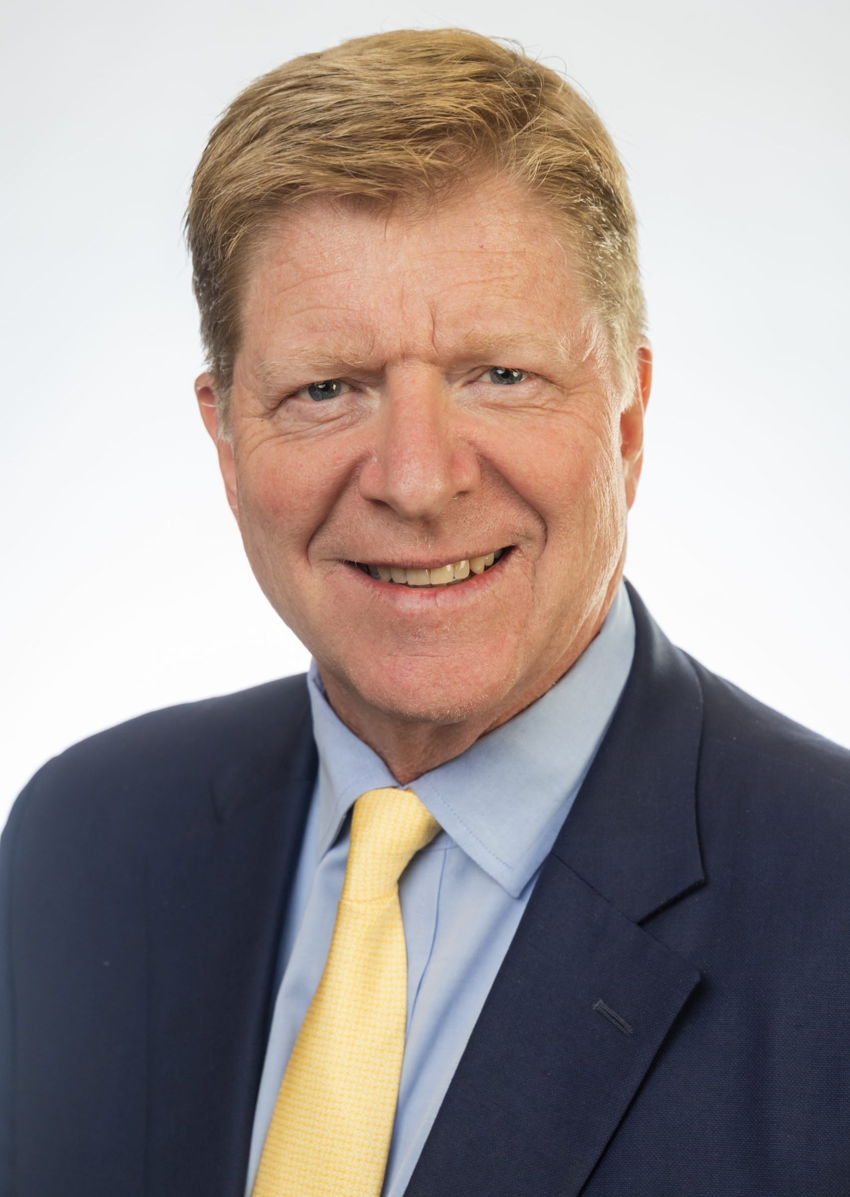 Headshot of a smiling man with light brown hair wearing a navy suit and yellow tie against a white background