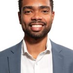 Headshot of a smiling man with short curly hair and a beard, wearing a light blue blazer and white shirt against a white background