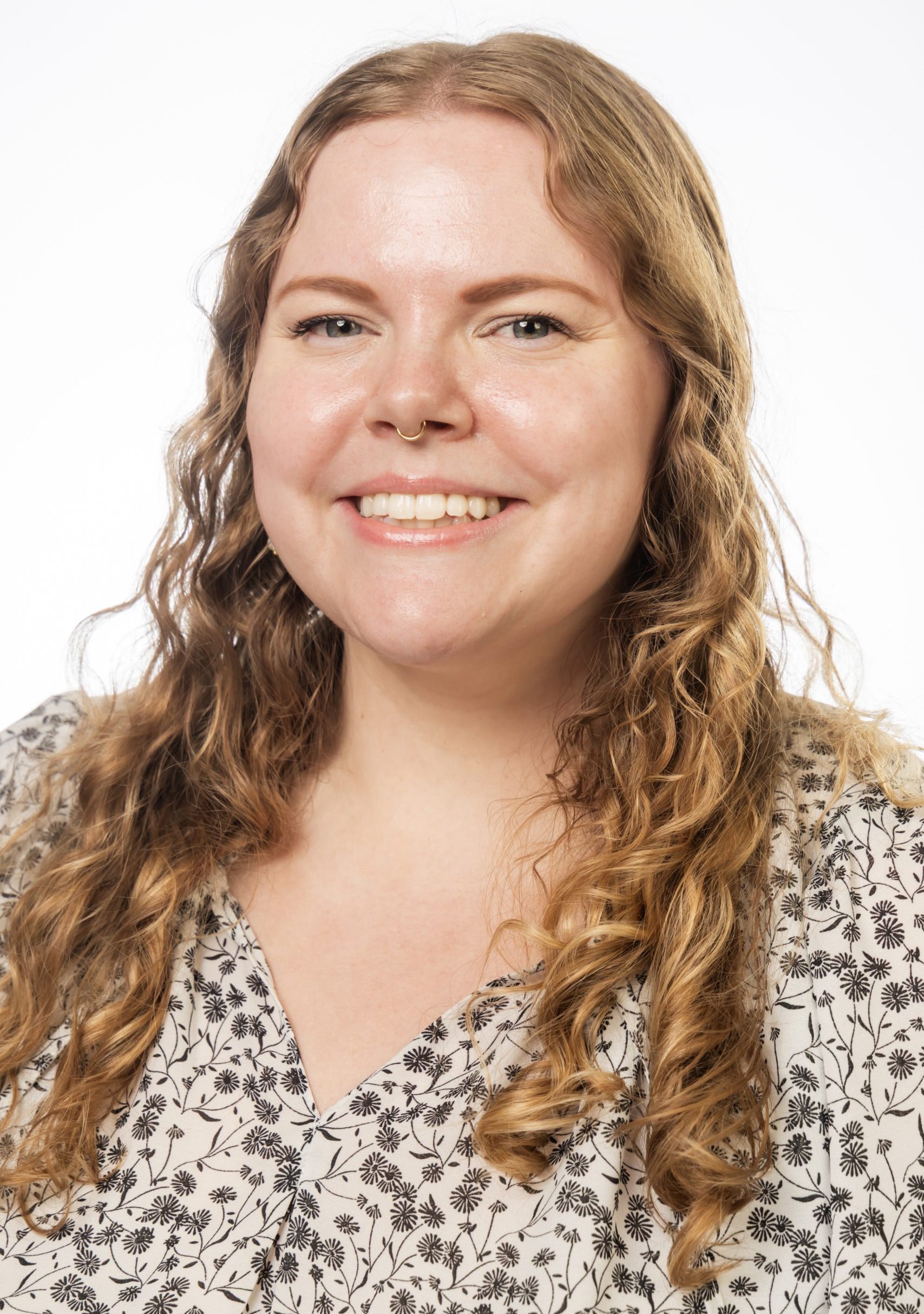 Headshot of a smiling woman with long curly light brown hair and a nose ring, wearing a patterned blouse against a white background