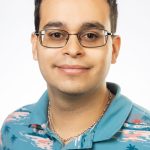Headshot of a man with short dark hair wearing glasses and a teal patterned collared shirt against a white background