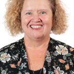 Headshot of a smiling woman with short curly blonde hair wearing a black floral top against a white background
