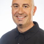 Headshot of a smiling bald man with facial hair wearing a black collared shirt against a white background