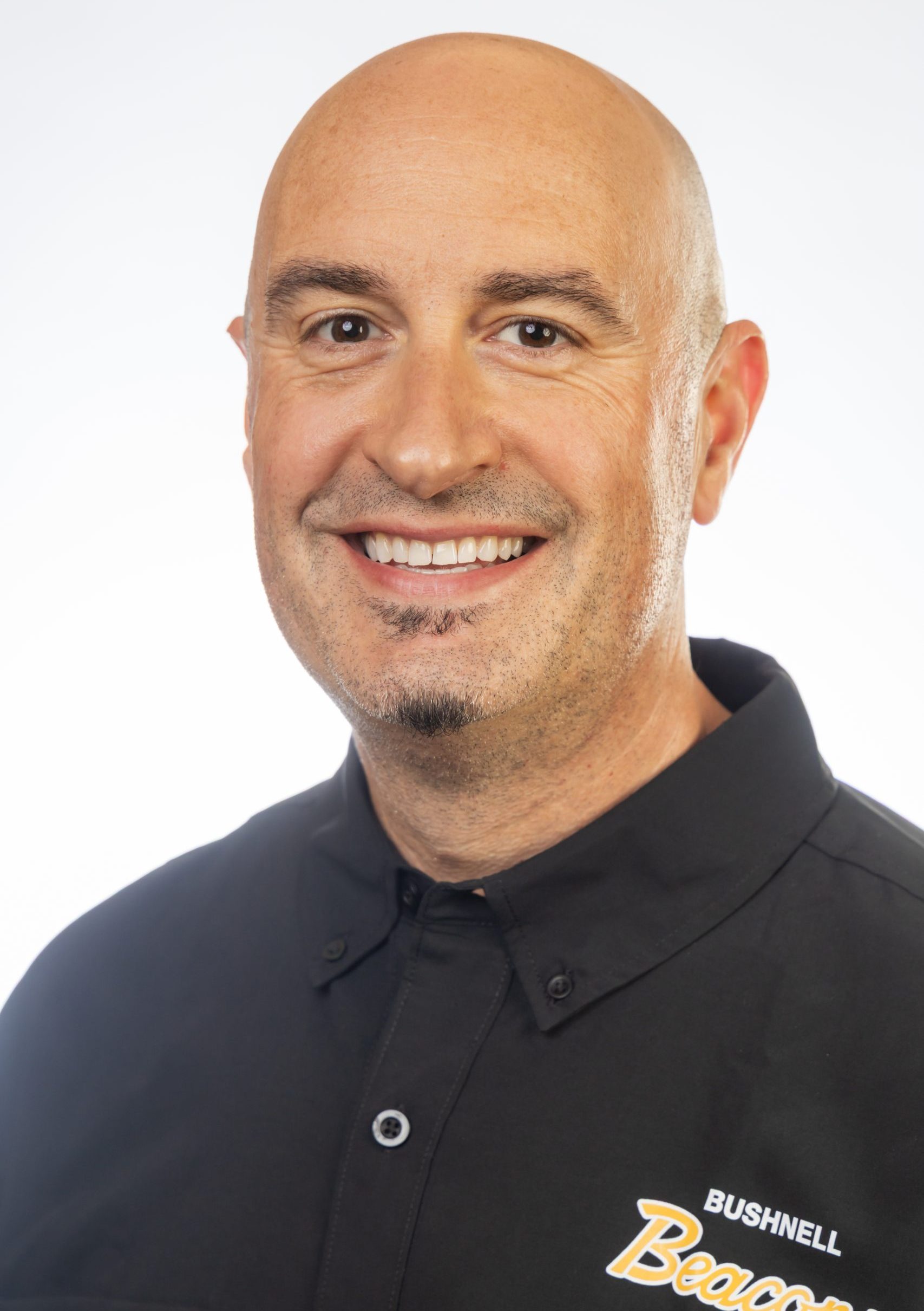 Headshot of a smiling bald man with facial hair wearing a black collared shirt against a white background