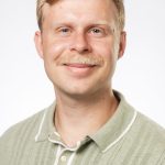 Headshot of a smiling man with short blonde hair and a mustache wearing a light green collared shirt against a white background