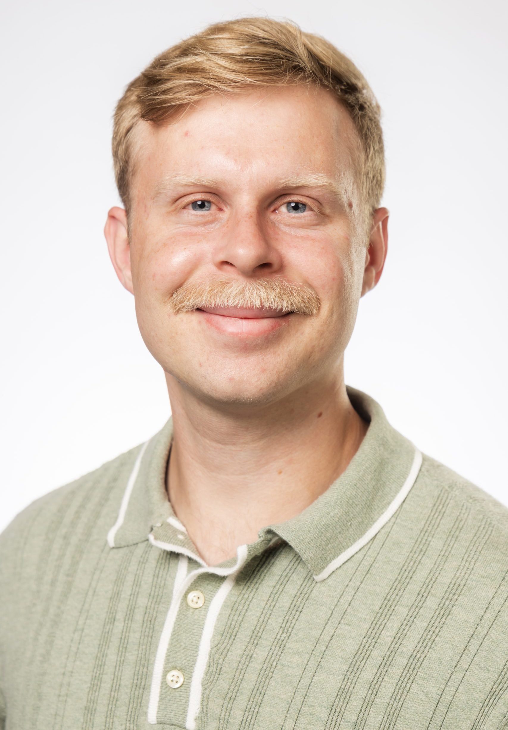 Headshot of a smiling man with short blonde hair and a mustache wearing a light green collared shirt against a white background