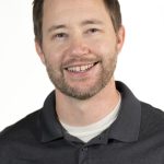 Headshot of a smiling man with short brown hair and a beard wearing a dark gray polo shirt against a white background