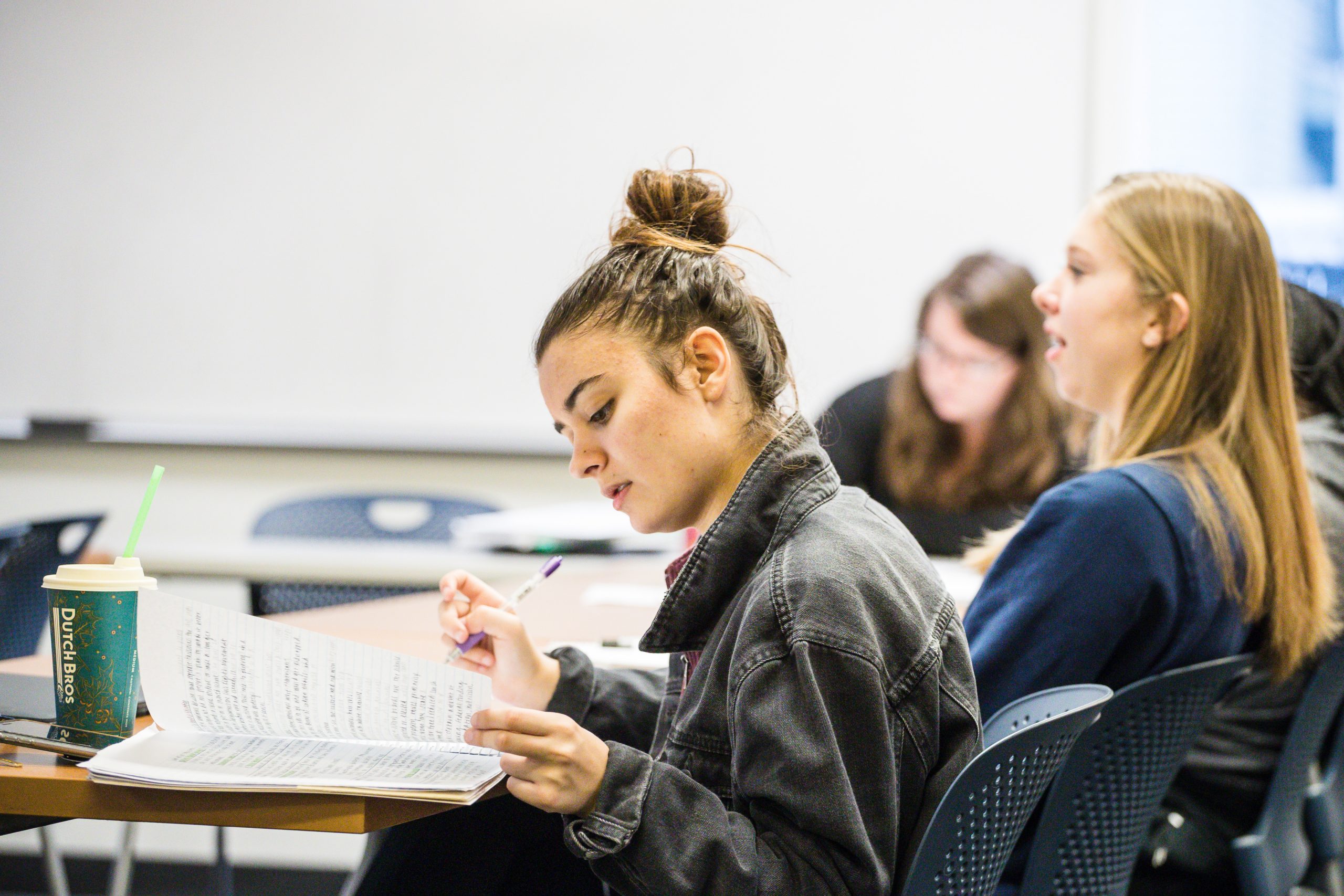 Student sitting at a classroom desk writing on a piece of paper with a pen