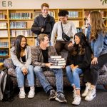 male and female students sitting on and around a couch in the library, stack of books on the lap of the middle student