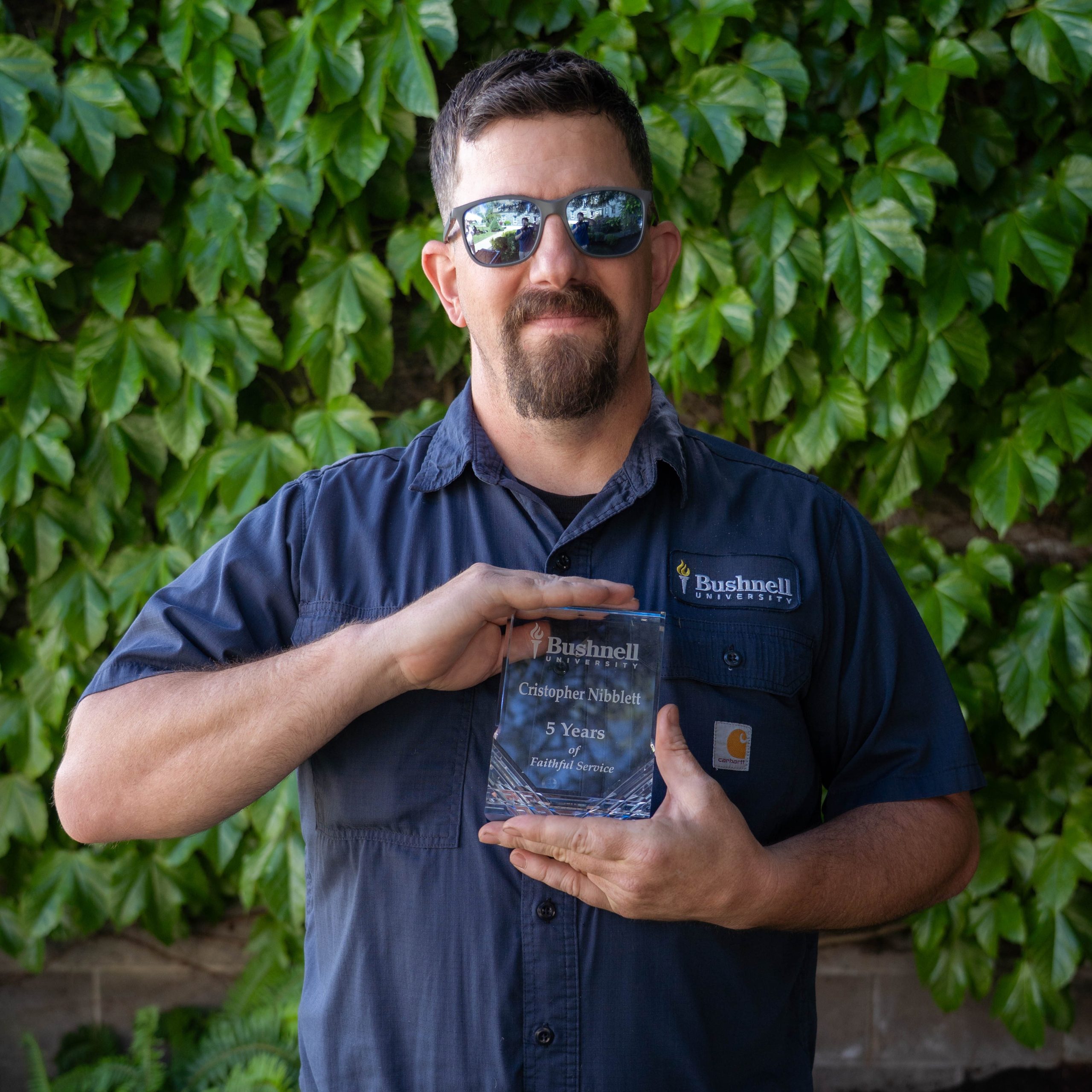 Employee recognized for 5 years of faithful service while holding a commemorative award outdoors on campus