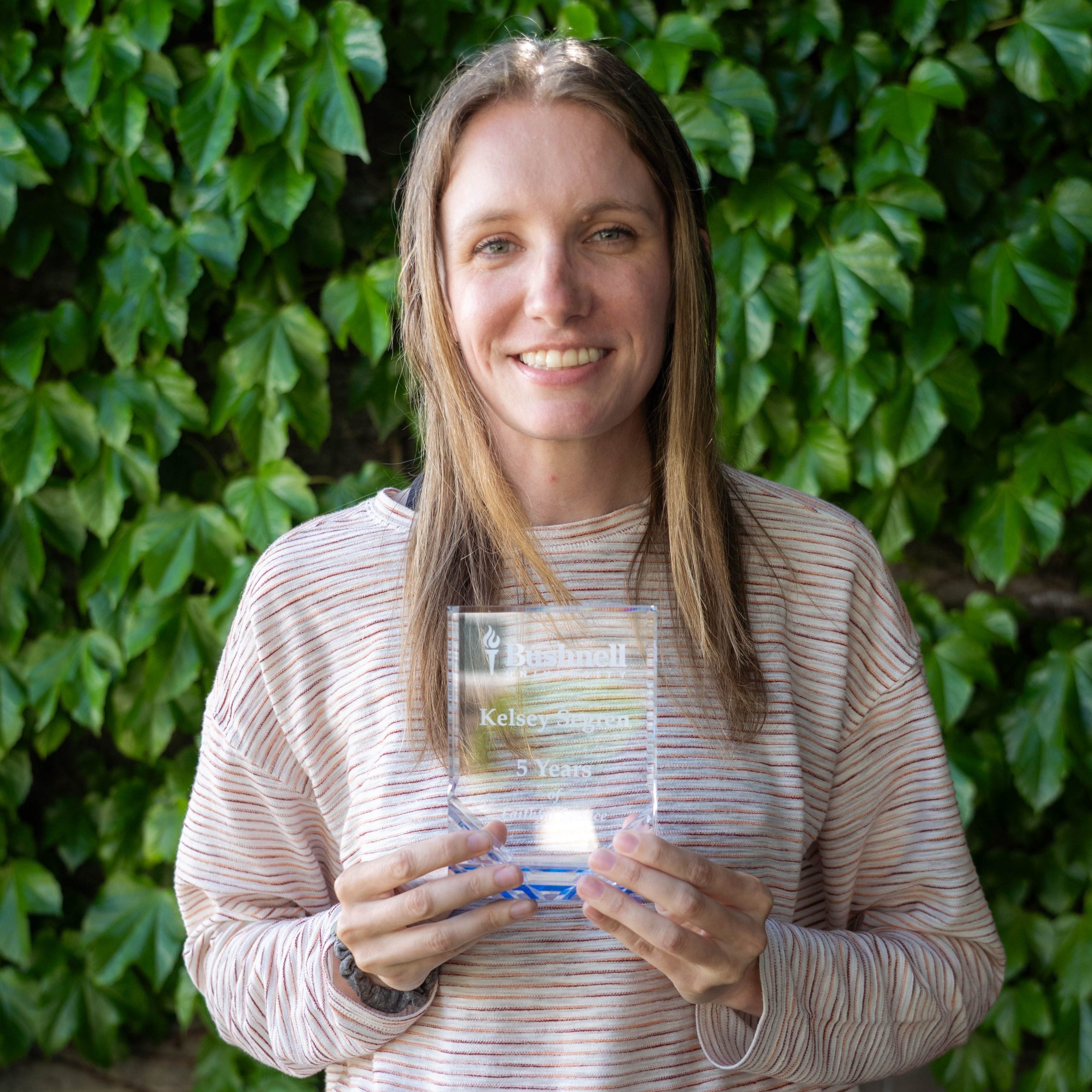 Employee recognized for 5 years of faithful service while holding a commemorative award outdoors on campus
