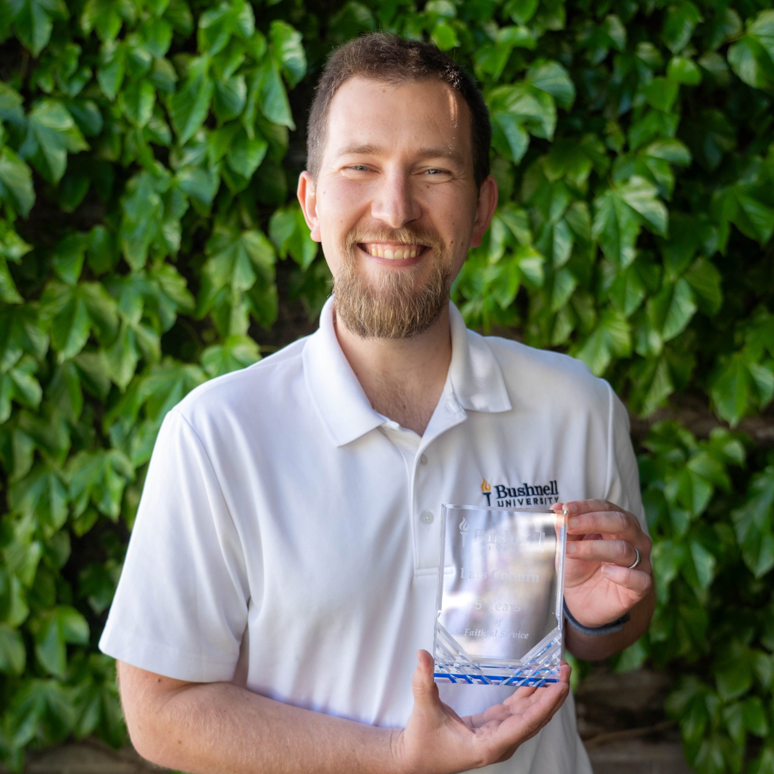 Employee recognized for 5 years of faithful service while holding a commemorative award outdoors on campus