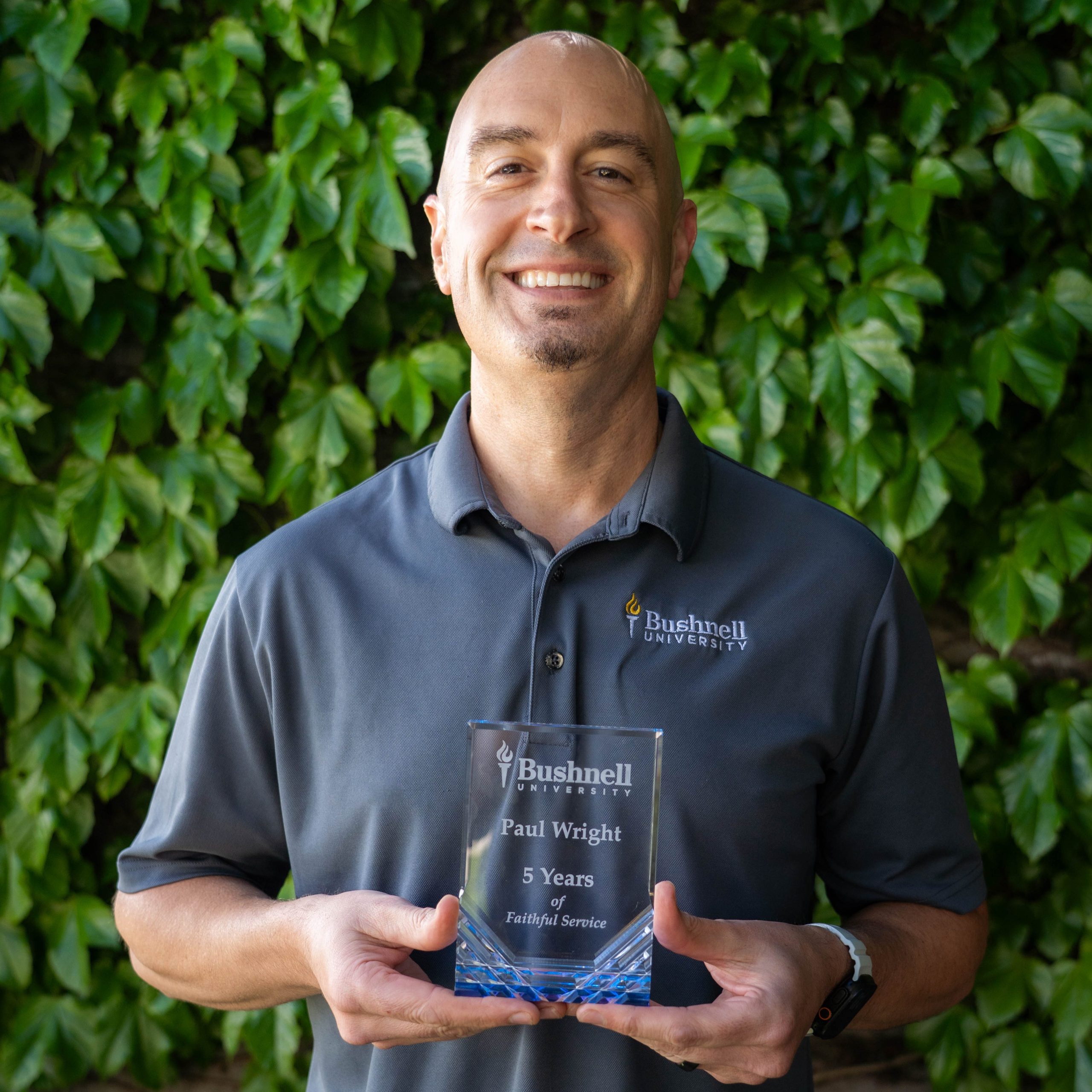 Employee recognized for 5 years of faithful service while holding a commemorative award outdoors on campus