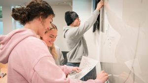 Three students collaborate at a classroom whiteboard, reviewing course materials and participating in an interactive learning activity.