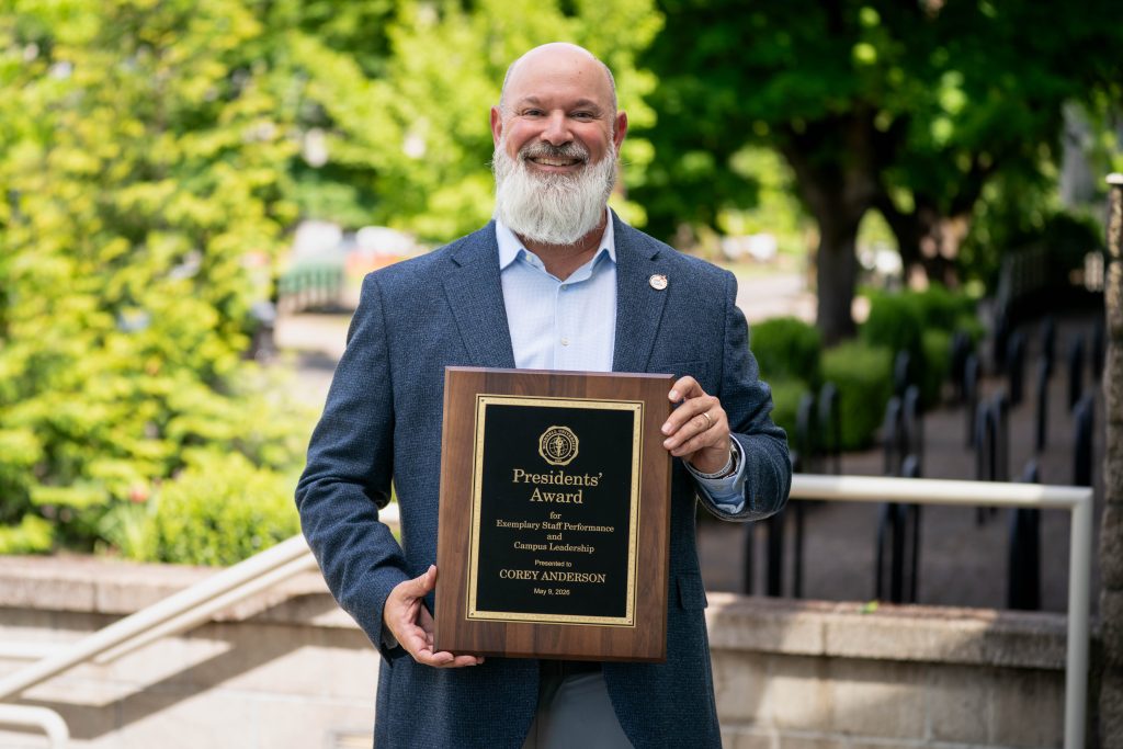 Staff member standing outdoors on campus holding a President’s Award plaque for exemplary staff performance and campus leadership