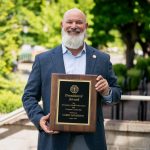 Staff member standing outdoors on campus holding a President’s Award plaque for exemplary staff performance and campus leadership