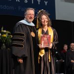 University president standing with a graduate in cap and gown holding an award plaque during a graduation ceremony