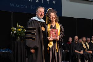 University president standing with a graduate in cap and gown holding an award plaque during a graduation ceremony