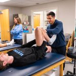 Athletic trainer aiding a student-athlete doing a leg stretch in a training facility