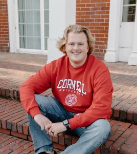 Male student sitting on the steps of brick building on campus with a red Cornel University sweater