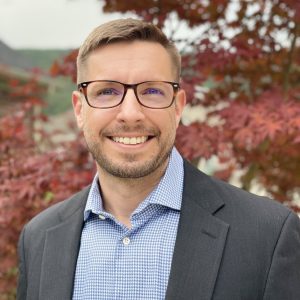 Professional headshot of a male businessman with glasses in a grey suit jacket with maple trees in the background