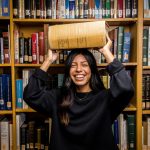 English student holding a large dictionary in a library surrounded by bookshelves