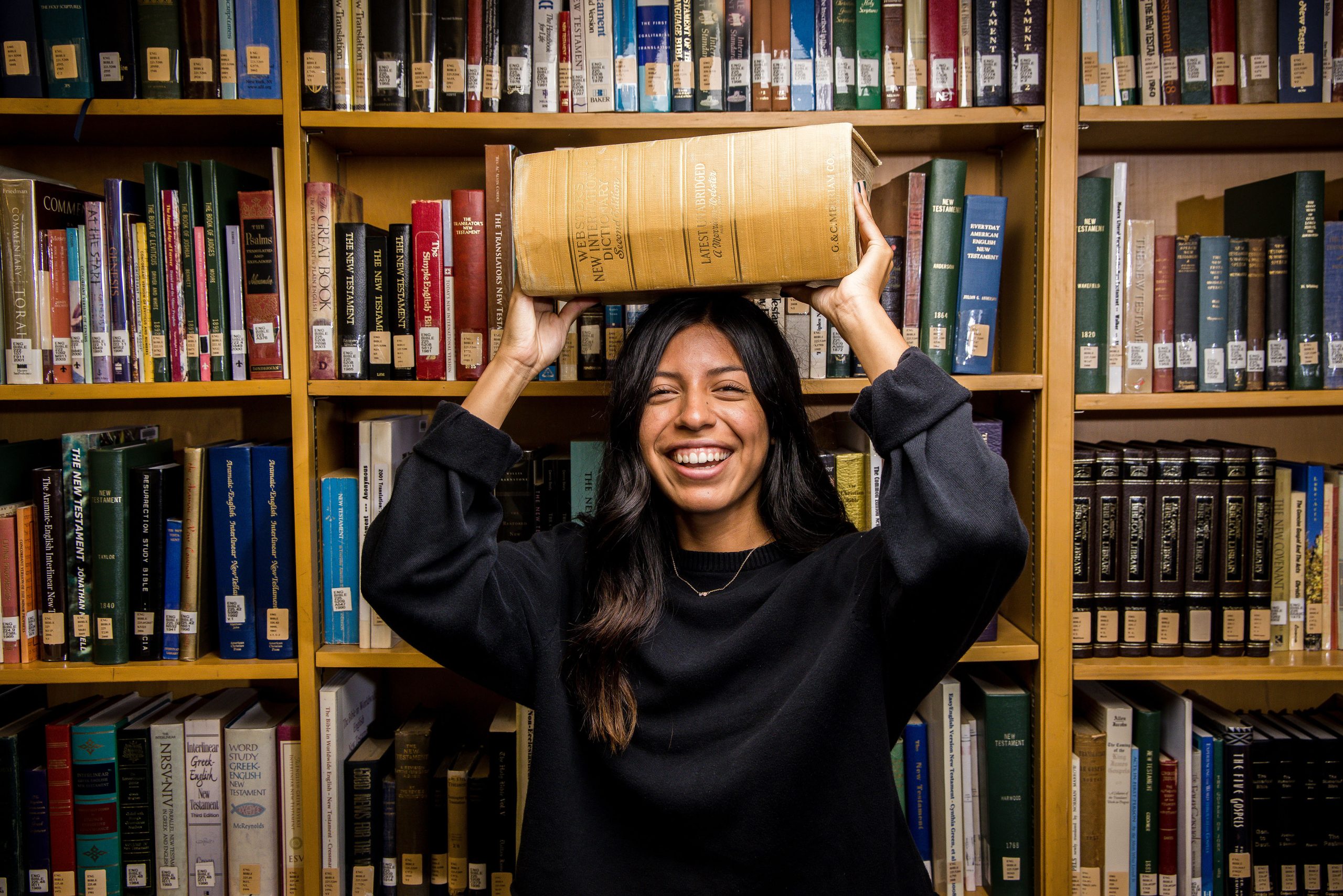 English student holding a large dictionary in a library surrounded by bookshelves