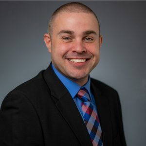 A confident professional headshot of an adult male in a black business suit and shaved head