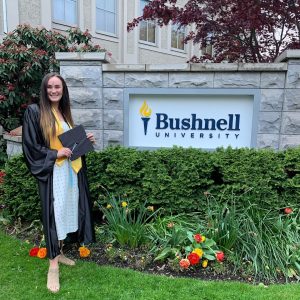 A education student in graduation cap and gown smiling in front of a Bushnell University monument sign