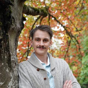 Student with a mustache and sweater headshot with a fall tree background