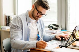 A young man working in a business office referencing his laptop while he writes on a notepad