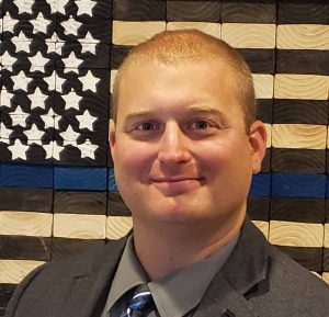 Headshot of a male police officer with shaved hair with a blue line flag behind
