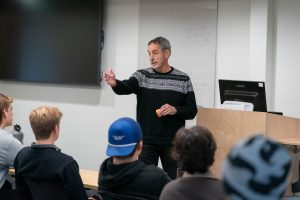 A male professor gives lecture at the front of a classroom with a black sweater