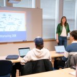 Female professor teaching at the front of a classroom with students using laptops