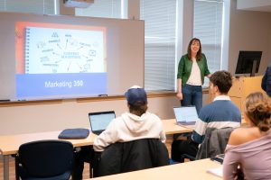 Female professor teaching at the front of a classroom with students using laptops