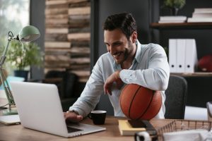 A professionally dressed sports team manager working on a laptop in their office while holding a basketball