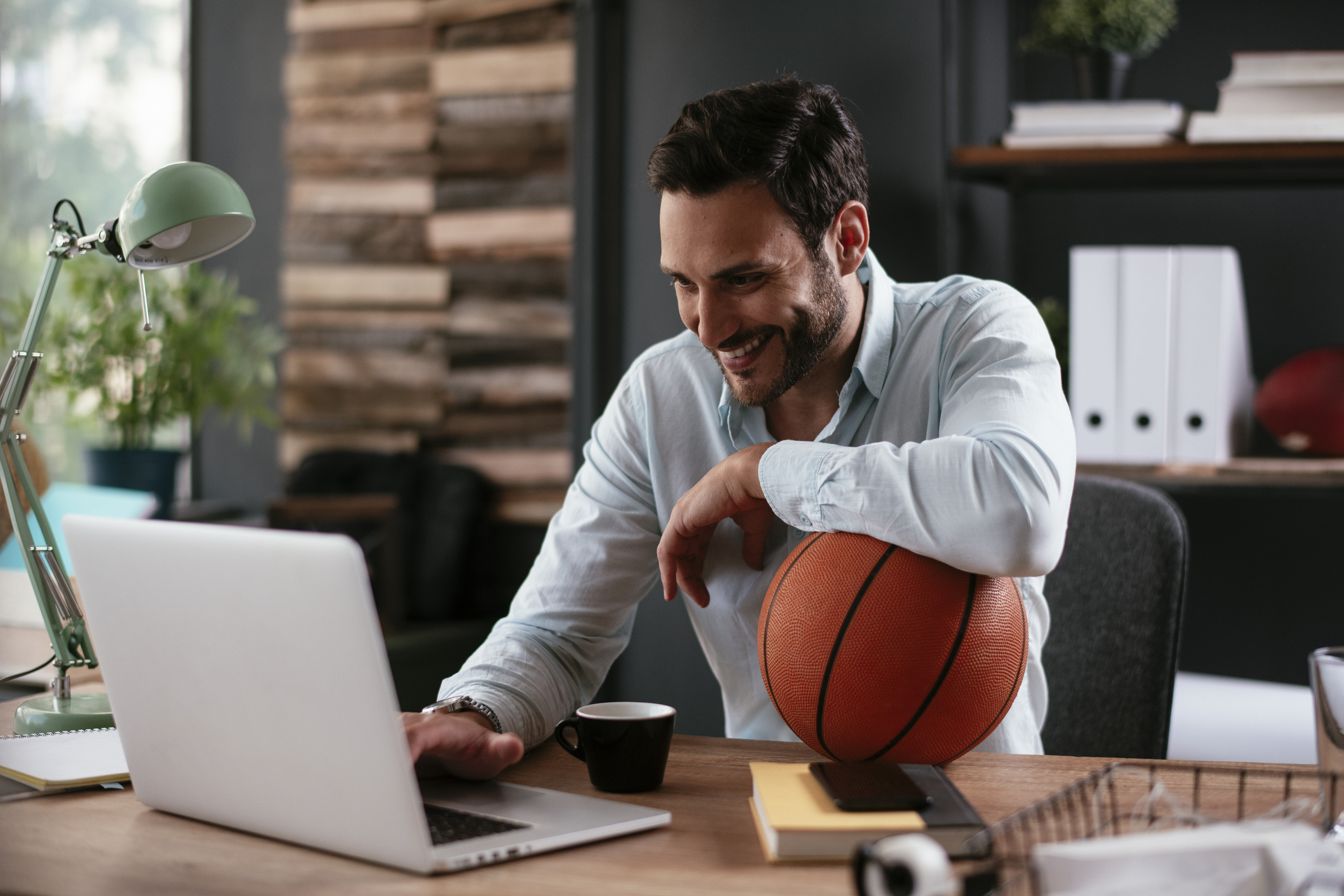 A professionally dressed sports team manager working on a laptop in their office while holding a basketball