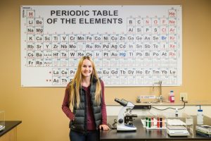 student standing in biology lab with microscope and periodic table of elements poster
