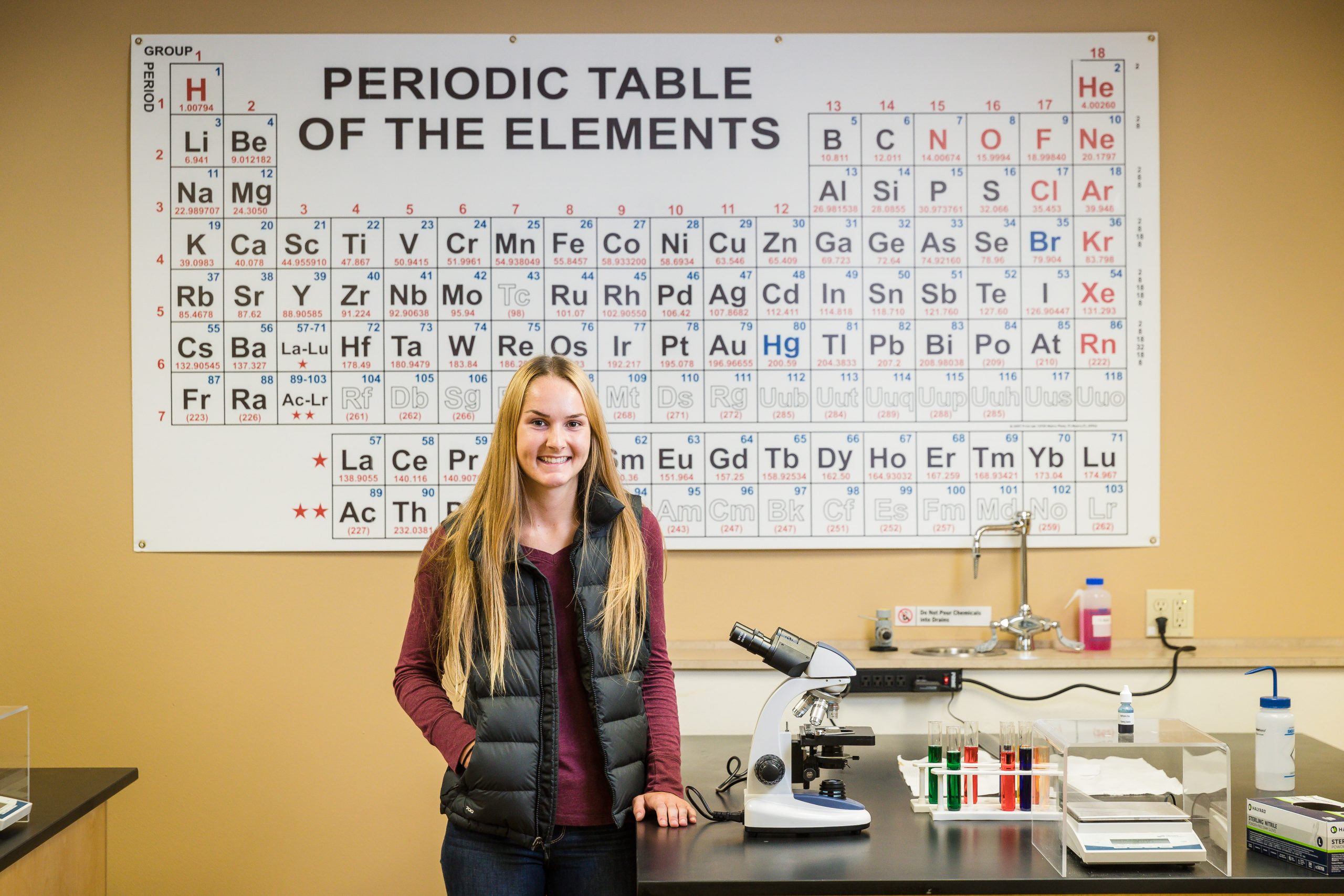 student standing in biology lab with microscope and periodic table of elements poster